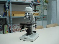 Microscopio ottico bianco su tavolo; sullo sfondo degli scaffali contenenti libri 